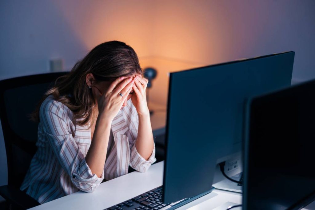 Tired and stressed out woman working late in an office, leaning at her desk and holding head in hands; anxious woman working overtime