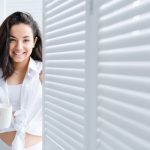 Modern young caucasian girl standing near the window with a cup of tea in the bedroom in the morning