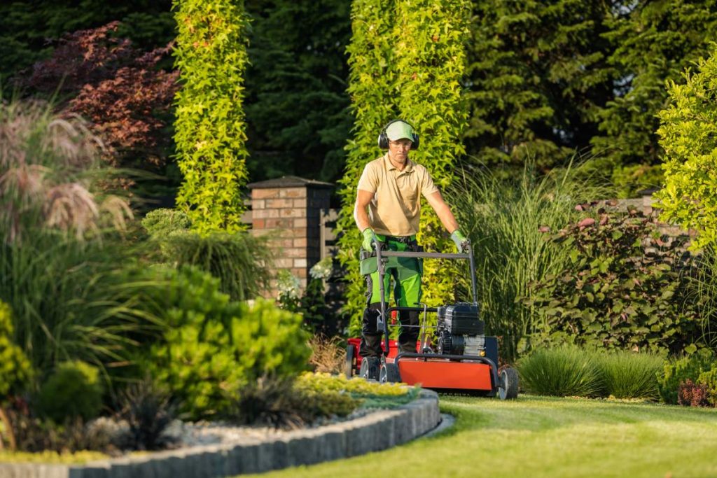 Large and Beautiful Landscape Garden in the Backyard of Residential House. Professional Landscaper in Protective Headphones Taking Care of Design Appearance by Mowing the Lawn.