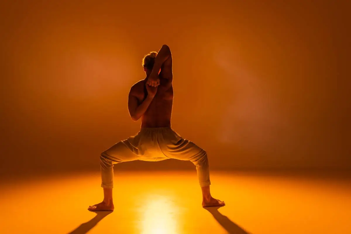 back view of shirtless man practicing goddess yoga pose with clenched hands behind back on orange