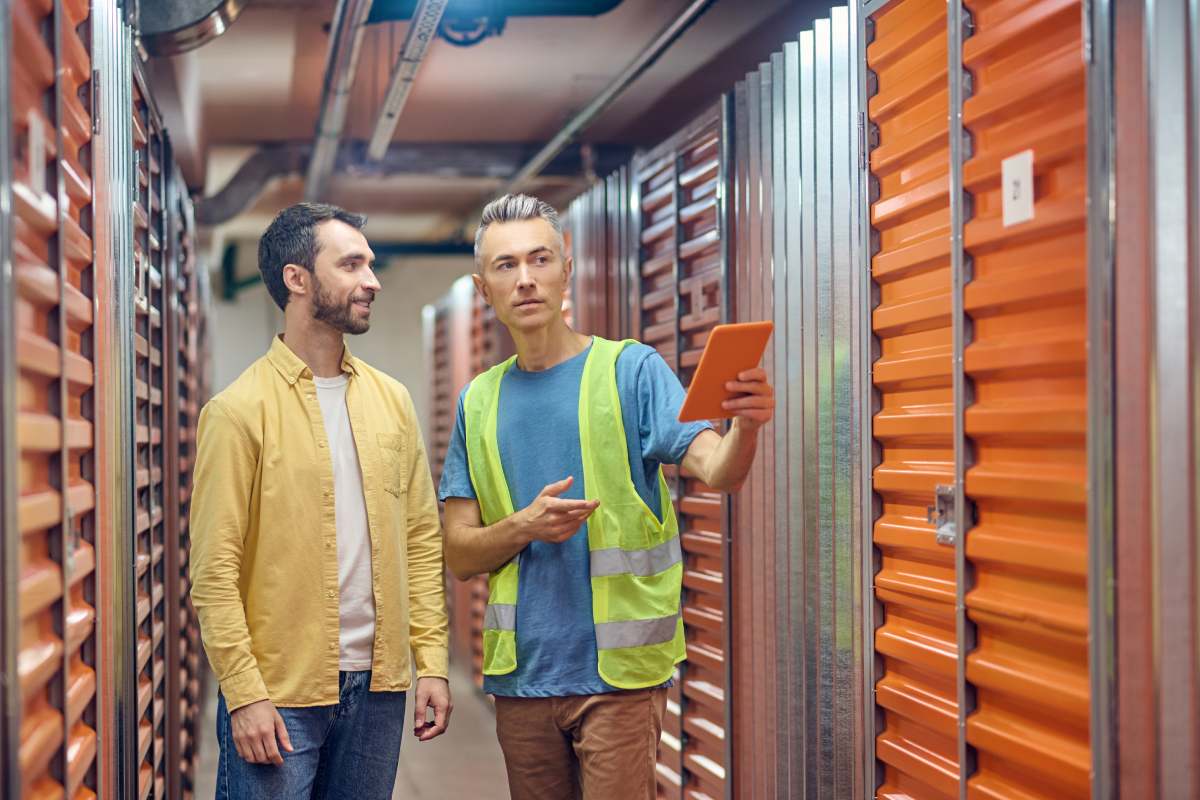 Warehouse, garage. Smiling young adult bearded shopper and warehouse worker in uniform with tablet pointing to number of nearest garage